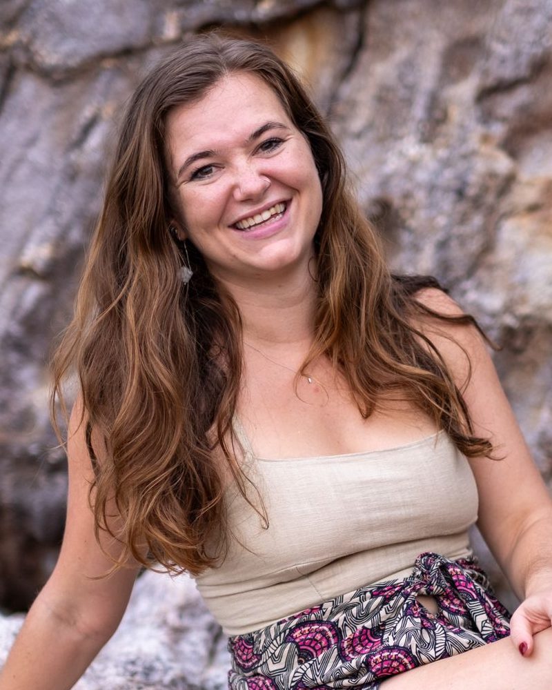 Mary Tichelaar, smiling, sitting against a rock face outdoors